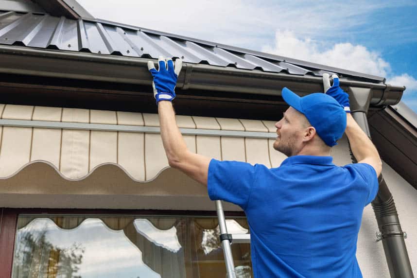 A man in a cap installing an aluminum gutter beneath metal roofing.