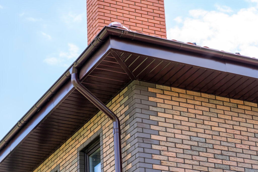 close-up view of house with brown roof and filing of roof overhangs with soffits and brick stone chimney pipe